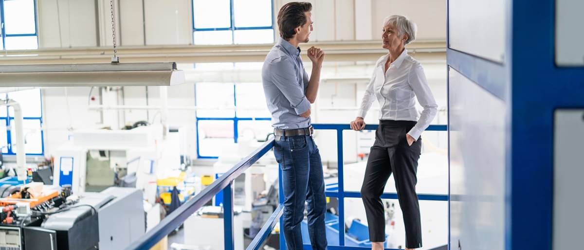 Jüngerer Geschäftsmann und ältere Geschäftsfrau sprechen in einer Fabrikhalle