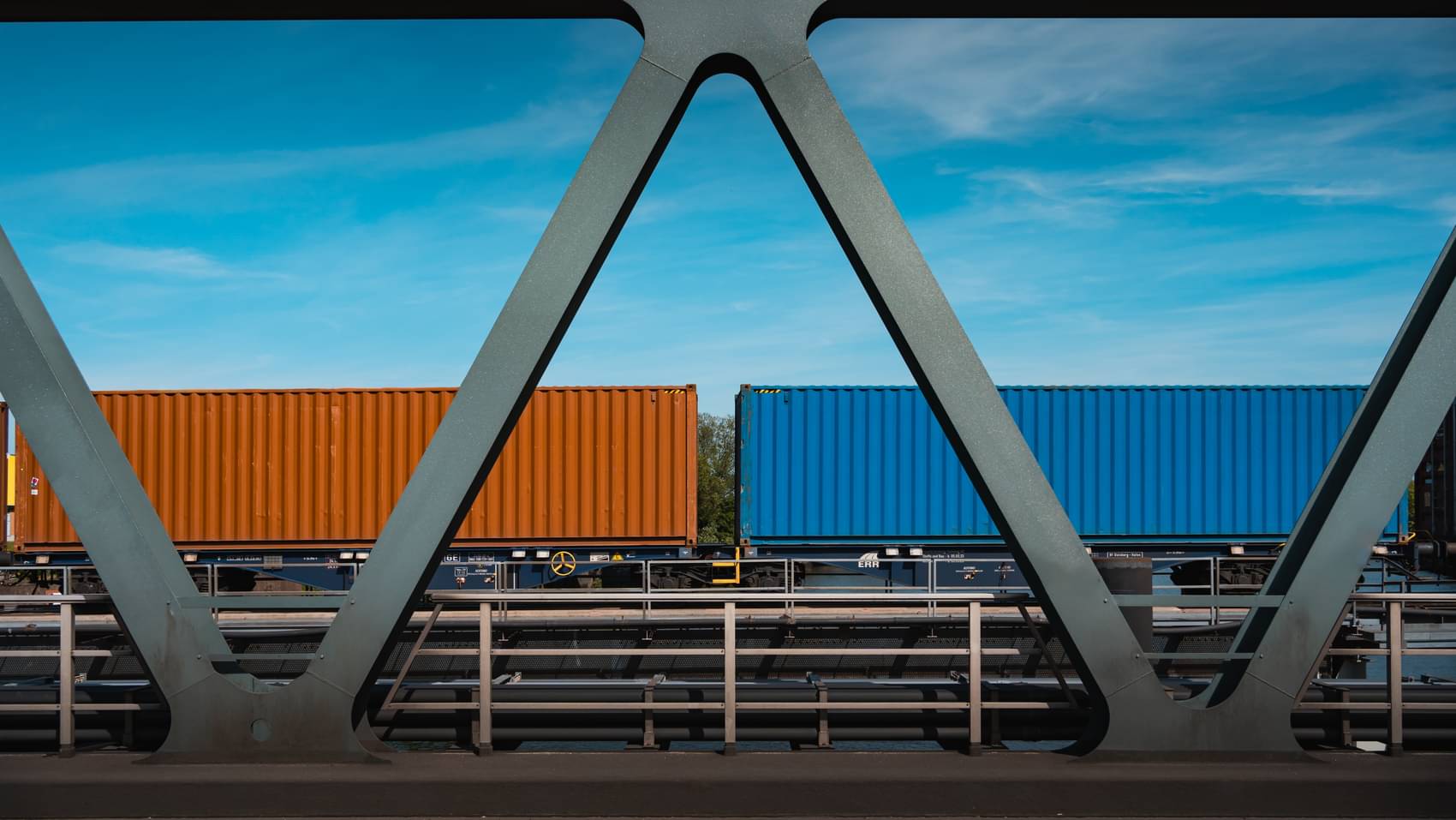 Aufnahme einer dunkel gefärbten Metallbrücke mit dreieckigem Fachwerkdesign, auf der ein Containerzug mit orangefarbenen und blauen Containern zu sehen ist.