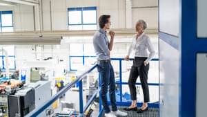 Jüngerer Geschäftsmann und ältere Geschäftsfrau sprechen in einer Fabrikhalle