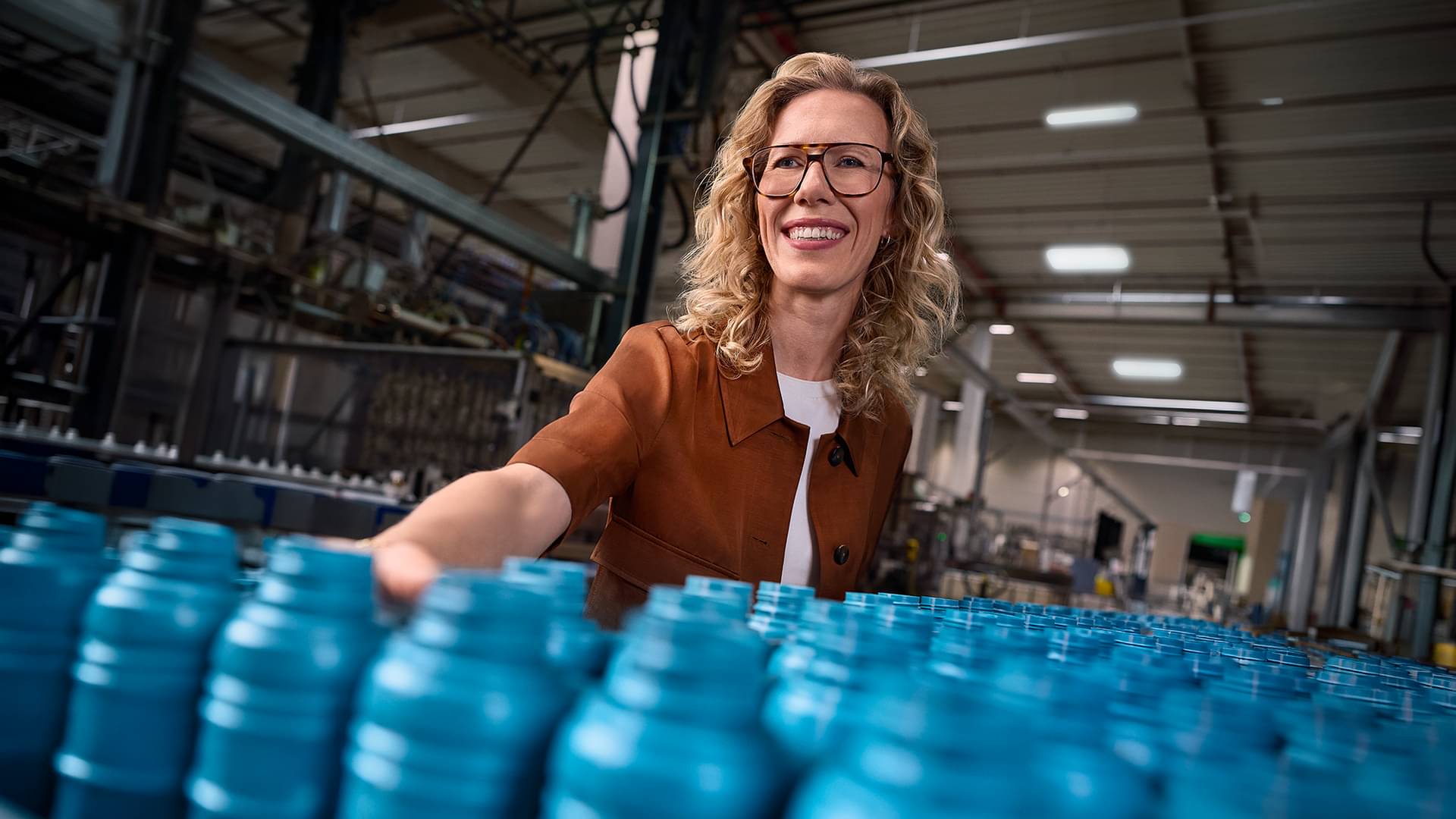 In a production hall, Dr. Alexandra Kohlmann, managing partner of ROWE, stands in front of a bottling line on whose conveyor belt blue bottles are transported.