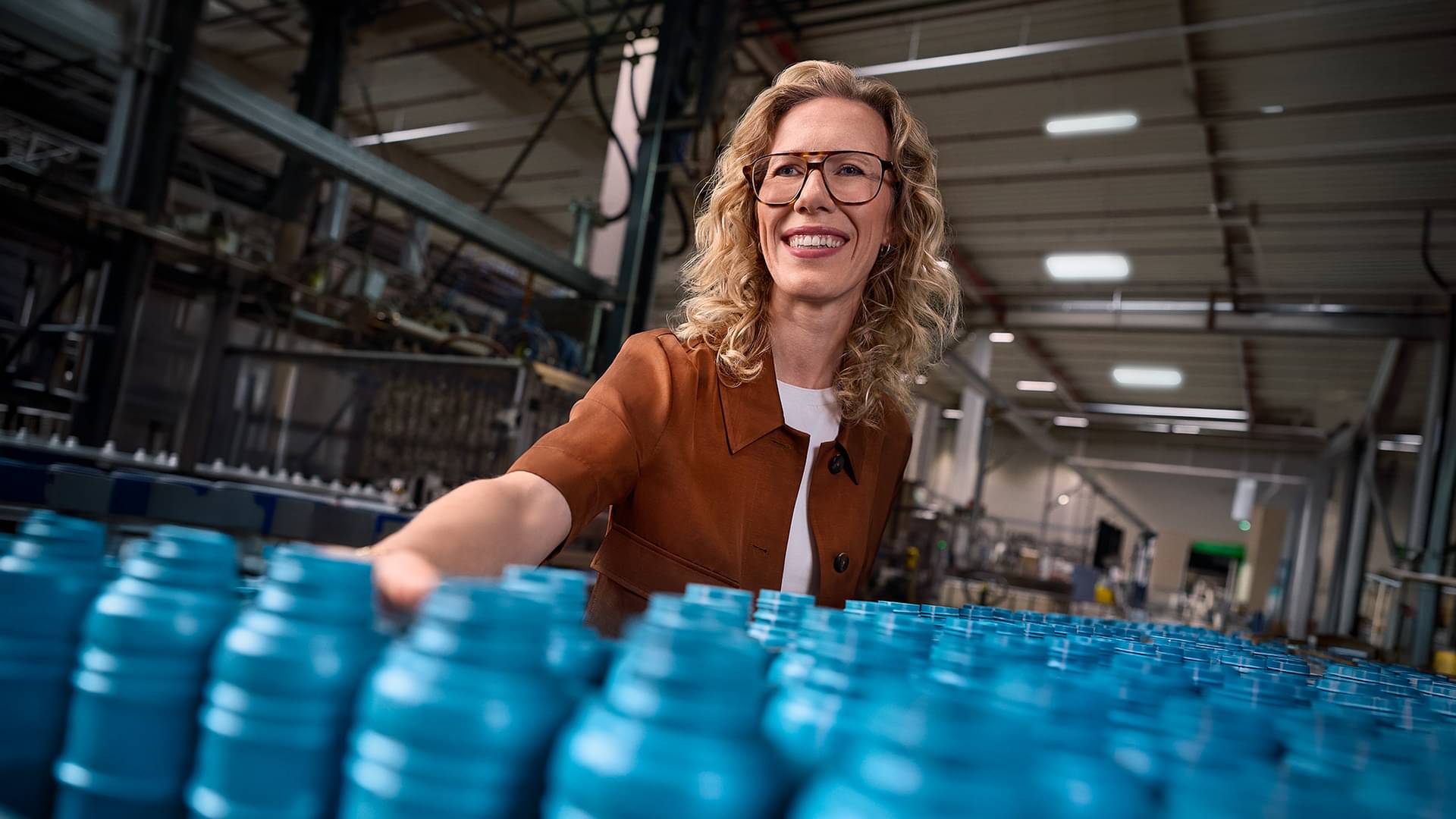 In einer Produktionshalle steht Dr. Alexandra Kohlmann, geschäftsführende Gesellschafterin von ROWE, vor einer Abfüllanlage, auf deren Laufband blaue Flaschen transportiert werden.